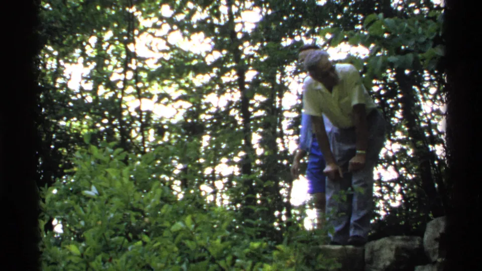 Adult man in casual clothing stands among rocks and trees in Isle Royale forest footage from the early 1970s.