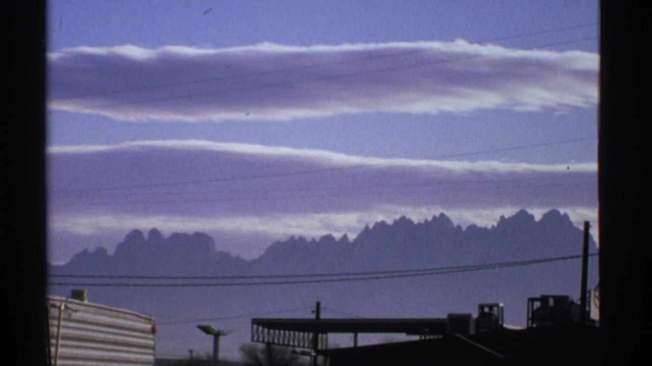 1972 New Mexico 8mm frame showing long bands of cloud above a distant mountain range, with dark buildings and wires in front.