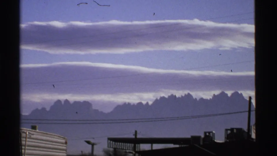 1972 New Mexico landscape frame with pale cloud layers, a jagged distant ridge, power lines, and dark foreground structures.