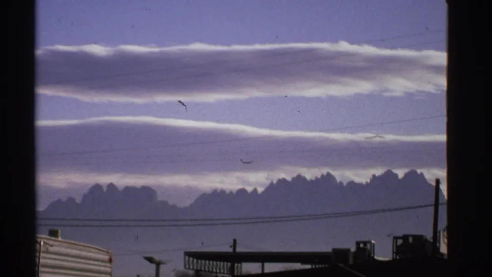1972 New Mexico 8mm frame with stacked cloud layers above distant mountains and dark roadside or industrial structures.