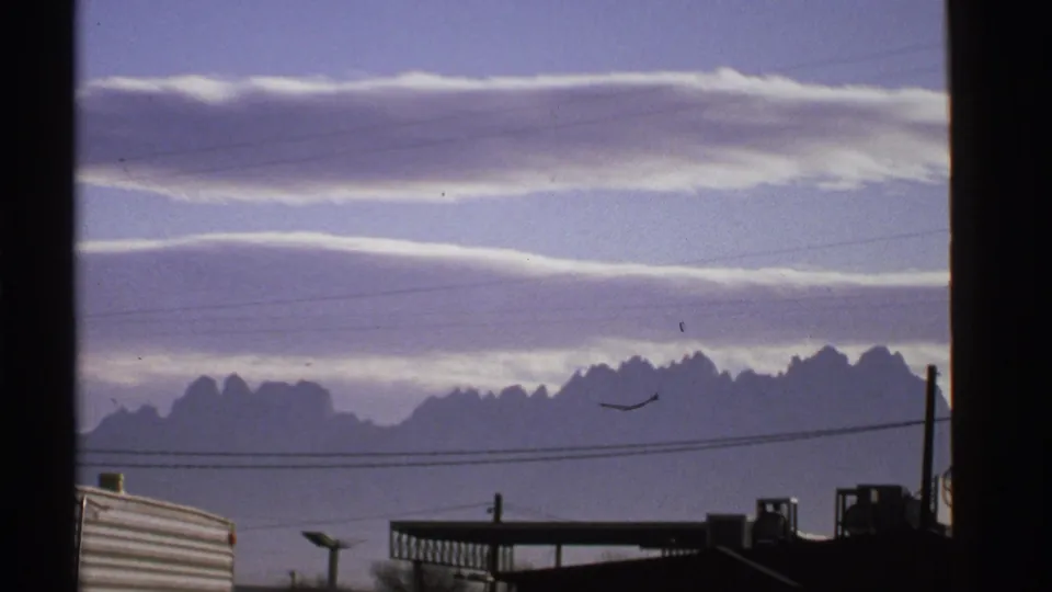 1972 New Mexico landscape frame showing cloud bands, distant mountains, foreground wires, dark buildings, and a small bird in the sky.
