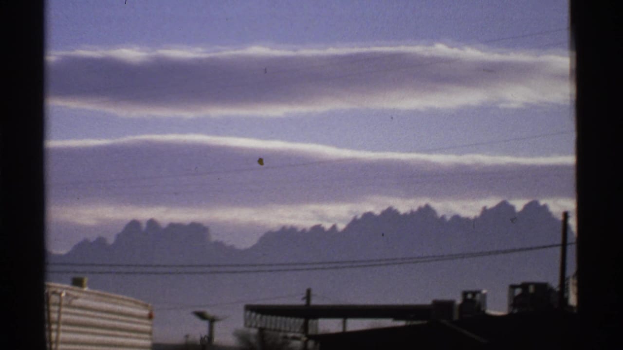 1972 New Mexico 8mm frame showing clouds over distant mountains, utility lines, and partial roadside structures before the facility sign appears.