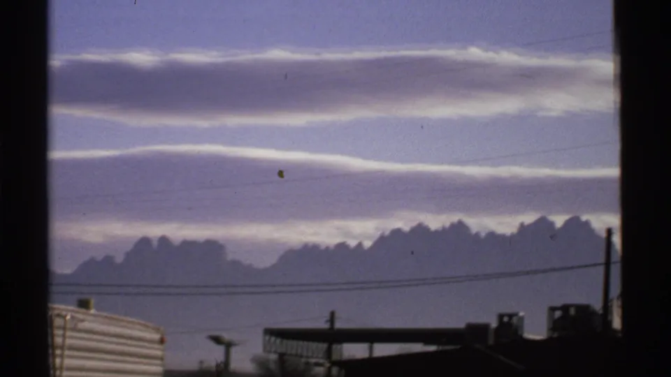 1972 New Mexico 8mm frame showing clouds over distant mountains, utility lines, and partial roadside structures before the facility sign appears.