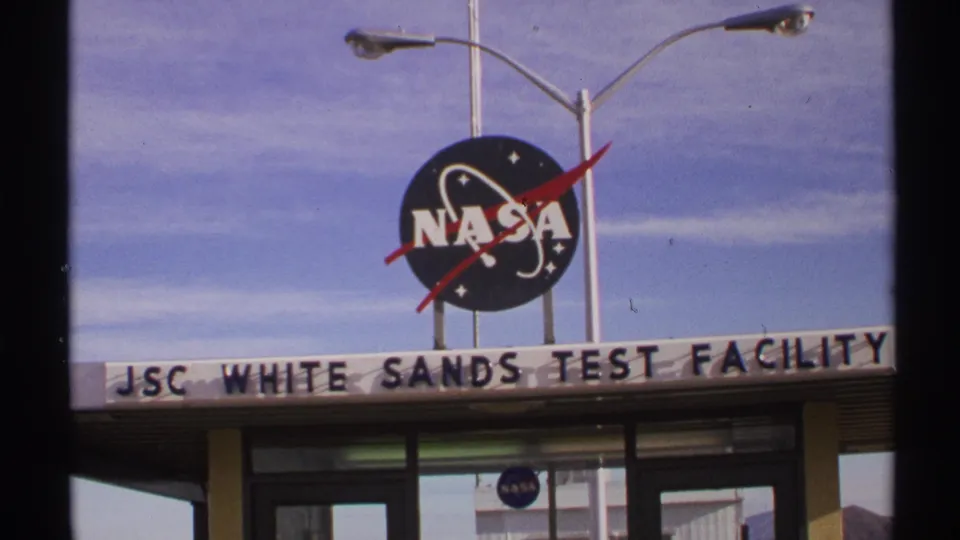 1972 New Mexico frame showing the NASA sign above JSC White Sands Test Facility lettering, streetlights, glass doors, and clear sky.