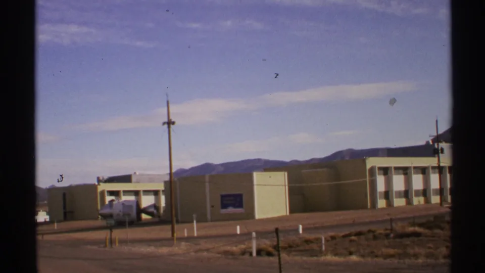 1972 New Mexico 8mm frame showing a beige roadside building, parked white vehicle, utility pole, desert ground, and distant mountains.