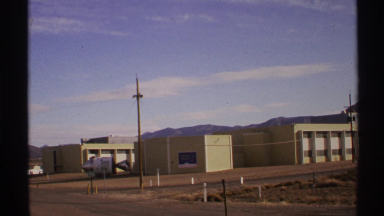 Roadside beige building with white-framed windows, utility pole, and mountains beyond.