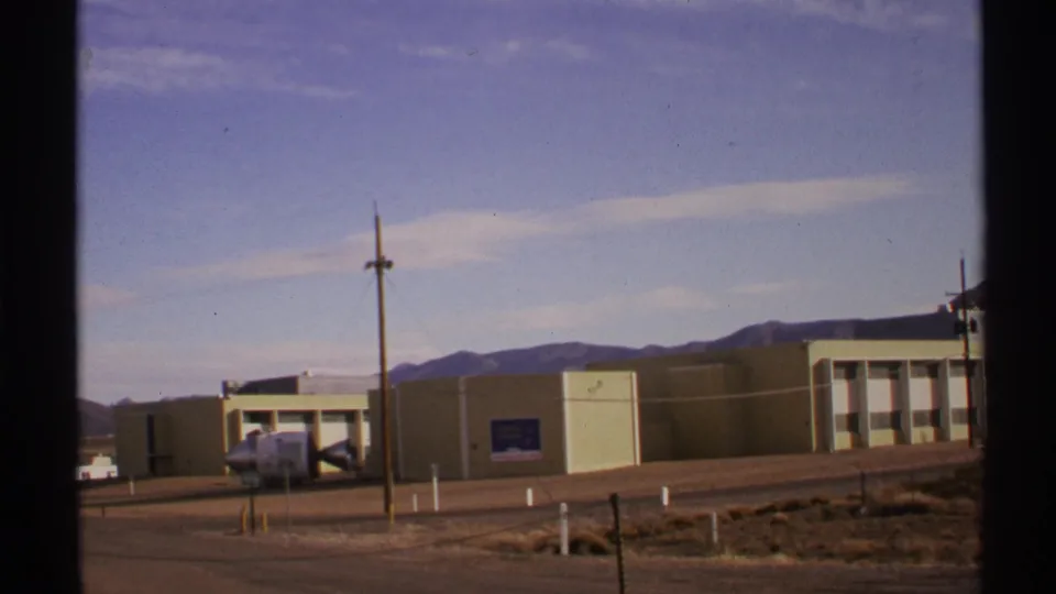 1972 New Mexico home-movie frame of a long beige building seen across a road, with a utility pole and low mountains in the distance.