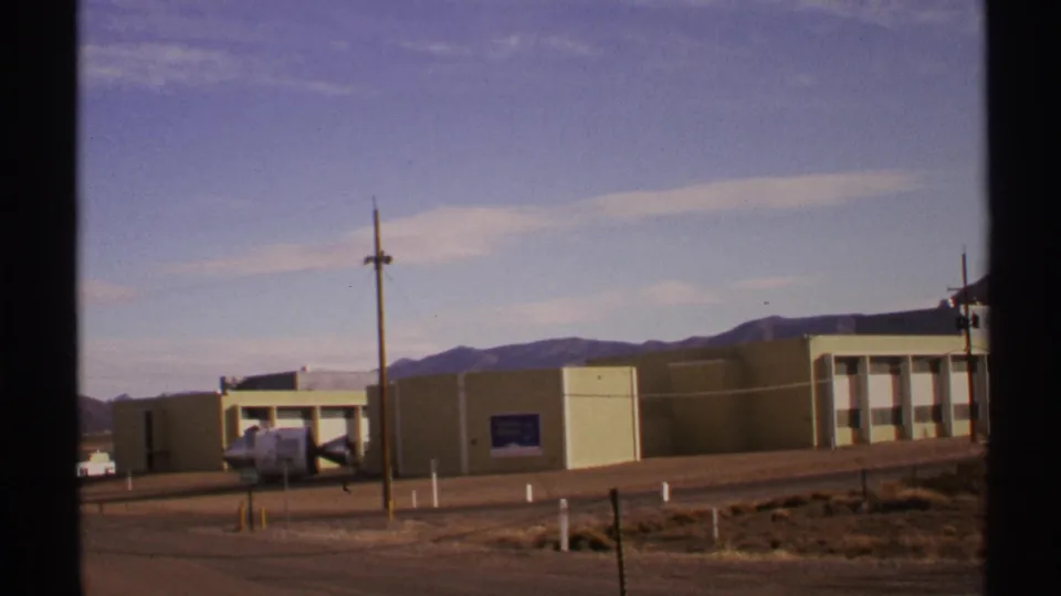 1972 New Mexico 8mm frame showing a beige building, a parked white vehicle, roadside posts, desert scrub, and a mountain backdrop.