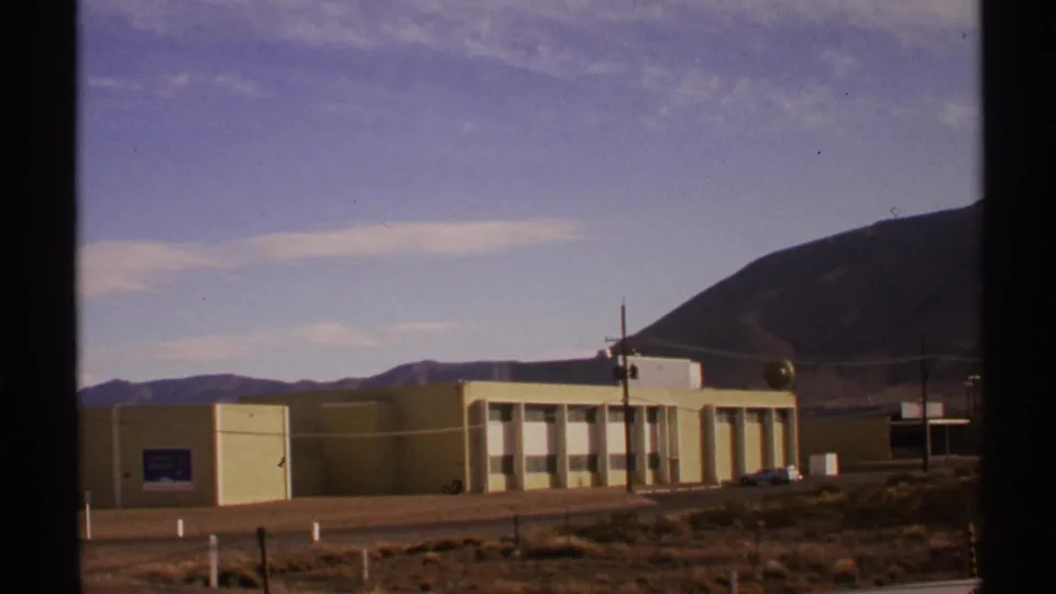 1972 New Mexico frame showing the building extending across the roadside, with desert ground, a utility pole, parked cars, and a large mountain behind.