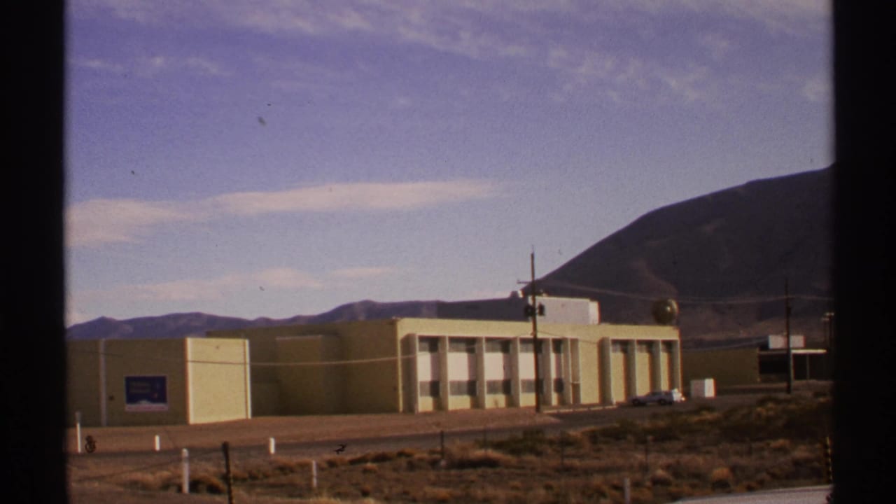1972 New Mexico 8mm frame showing a pale yellow modern building, desert scrub, utility poles, and hills in the background.