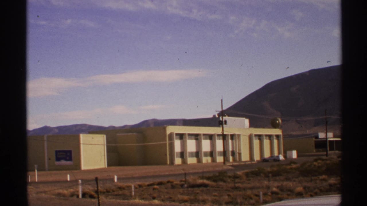 Exterior view of a yellow building with hills, poles, and a parked car nearby.