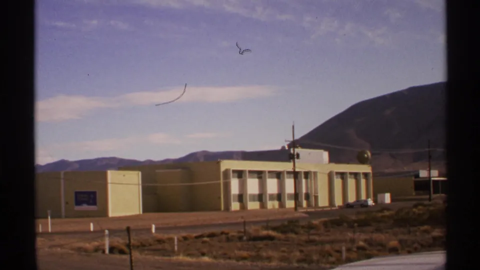 1972 New Mexico frame showing the institutional building across scrubby ground, with hills, poles, and film scratches visible.