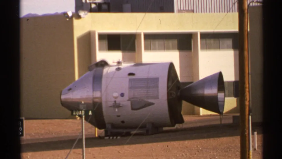 1972 New Mexico 8mm frame showing a silver Apollo-style capsule on an outdoor pad before a beige institutional building.