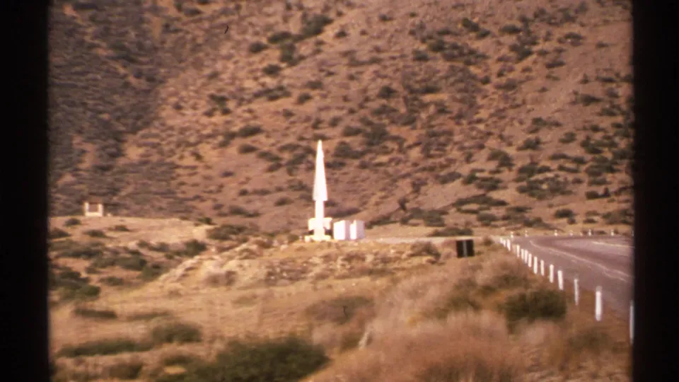 1972 New Mexico 8mm frame showing a distant white missile- or rocket-like structure near a road in a dry hillside landscape.