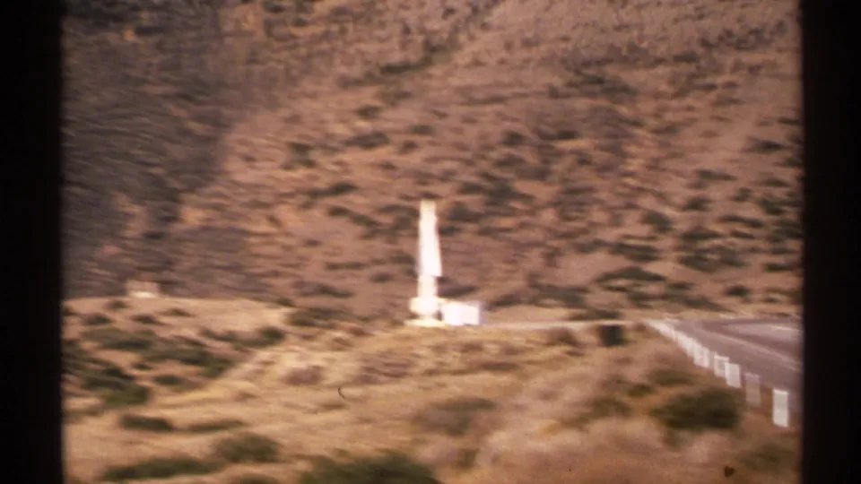 Motion-softened 1972 New Mexico home-movie frame with a white missile- or rocket-like structure near a road and scrub-covered hills.