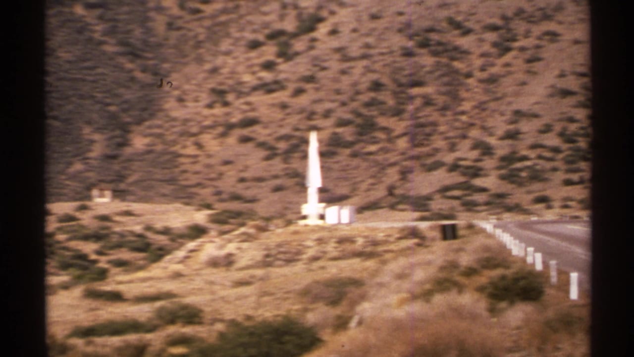 1972 New Mexico 8mm frame showing a white rocket-like object on a dry hillside near a road with white posts and rugged terrain.