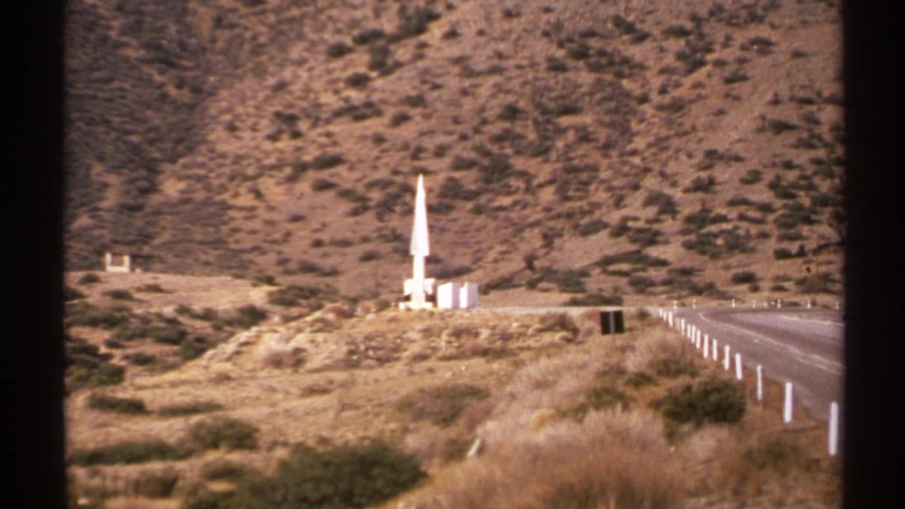 Roadside rocket-like object on a hill with guardrail posts and desert mountains behind it.