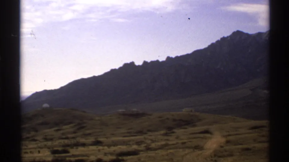 1972 New Mexico 8mm frame showing a rugged mountain ridge, rolling dry hills, and a small light structure in open country.