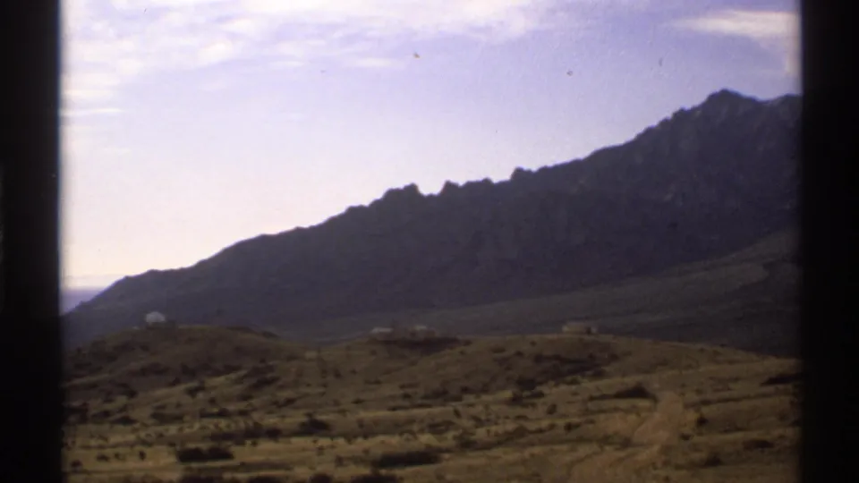 1972 New Mexico home-movie frame with a broad mountain ridge, dry foreground hills, sparse vegetation, and visible 8mm frame border.