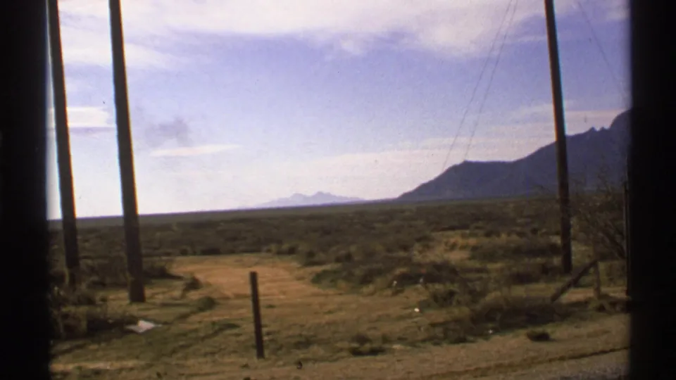 New Mexico desert road with utility poles, scrubland, distant mountains, and a pale blue sky in 1972 8mm footage.