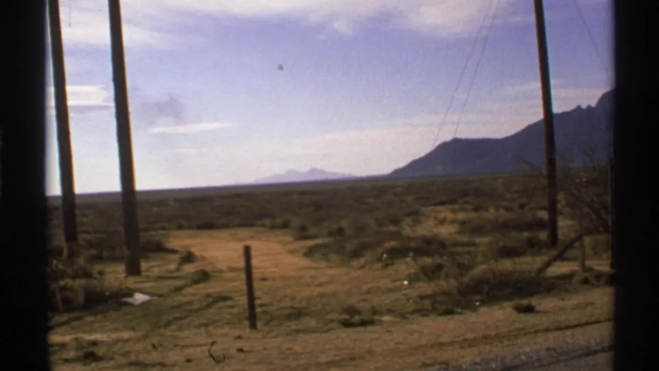 Passenger-window view of a desert road with utility poles, scrub vegetation, and distant mountain silhouettes in New Mexico.