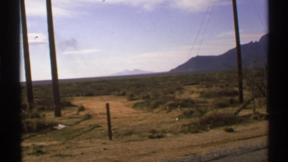1972 New Mexico desert view with a dirt road, utility poles, roadside scrub, and hazy mountains in the distance.