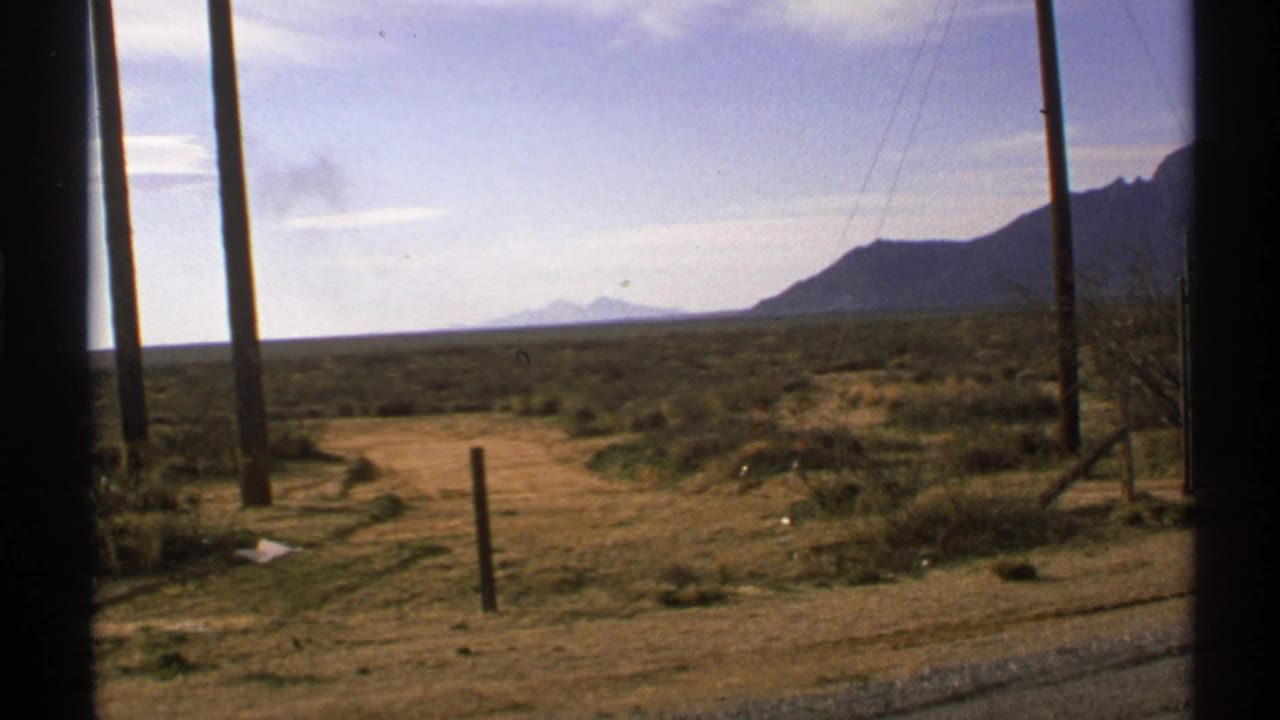 1972 New Mexico 8mm view across a scrubby roadside plain, framed by a vehicle window with poles and mountains in the distance.