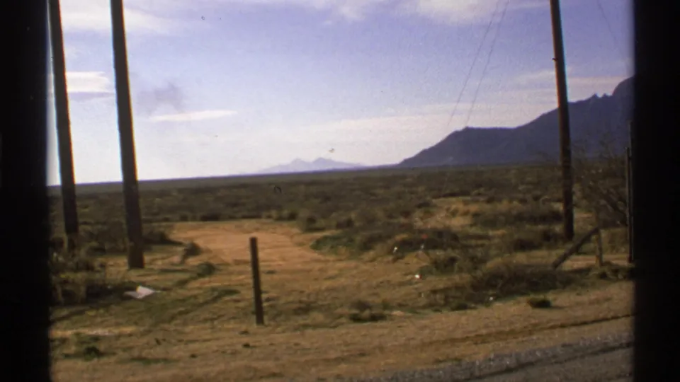 1972 New Mexico 8mm view across a scrubby roadside plain, framed by a vehicle window with poles and mountains in the distance.