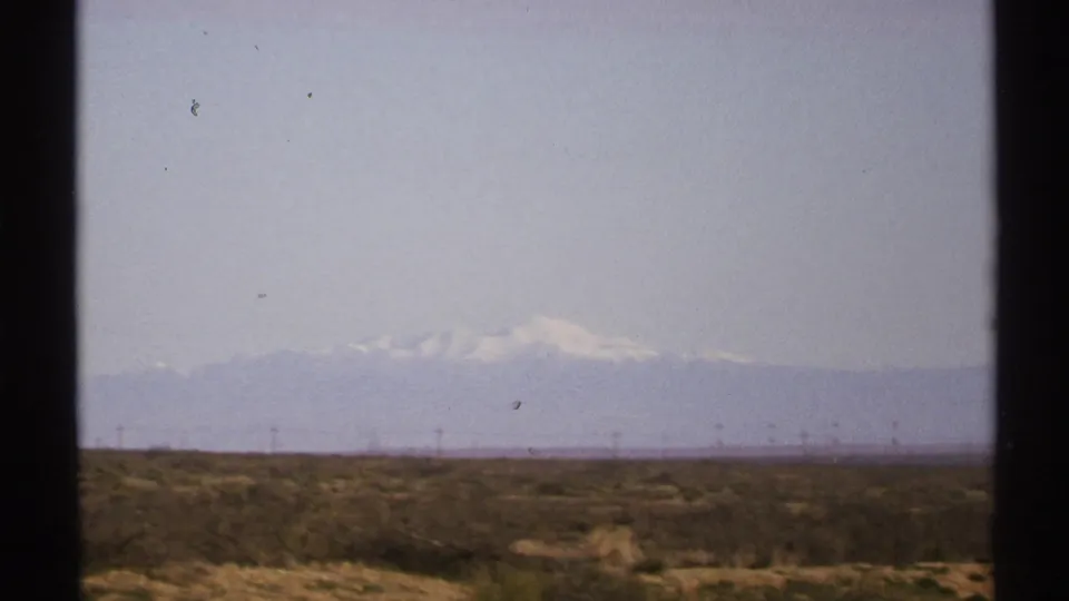 1972 New Mexico home-movie frame showing a flat desert plain, faint utility lines, and pale snow-capped mountains under a washed sky.