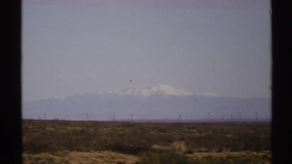 1972 New Mexico 8mm landscape view with scrub in the foreground, thin utility poles, and pale mountains softened by haze.
