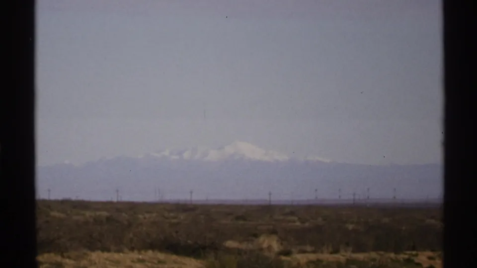 1972 New Mexico home-movie still of an open desert foreground, utility line silhouettes, and snow-capped mountains fading into the haze.