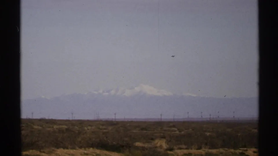 1972 New Mexico 8mm frame showing arid scrubland, utility poles, and distant snow-capped mountains under a pale sky.