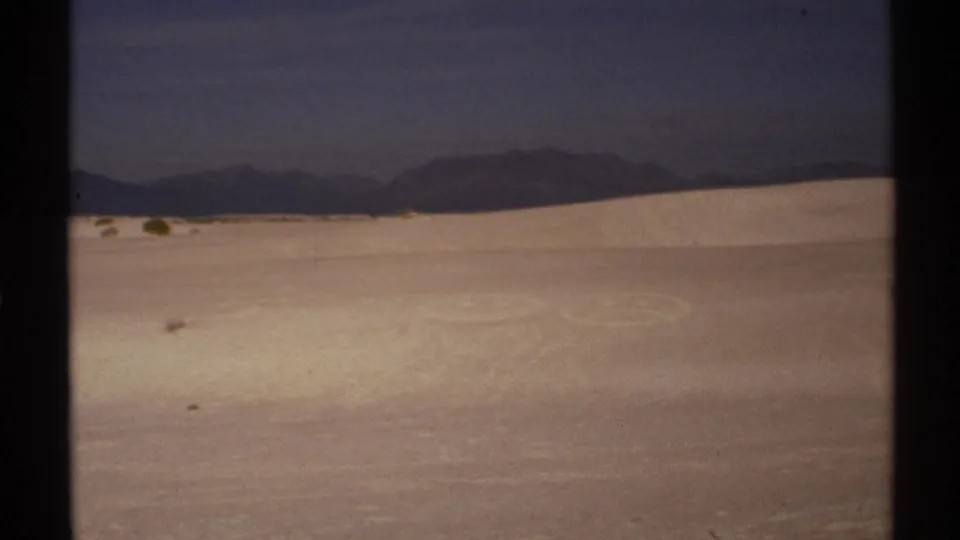 1972 New Mexico 8mm frame showing a broad white dune field, faint circular marks in the sand, and mountains on the horizon.