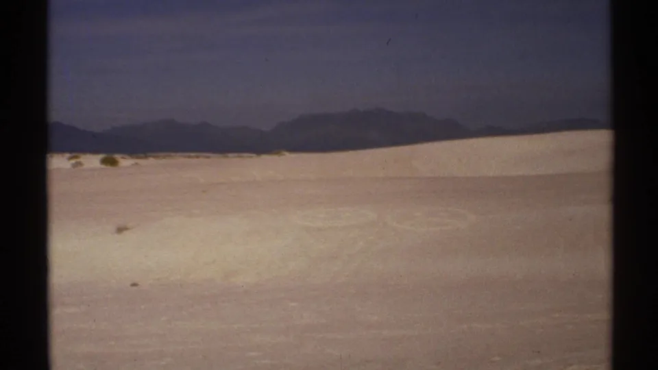 1972 New Mexico 8mm frame showing pale dunes with subtle tracks or circular marks and dark mountain silhouettes beyond.