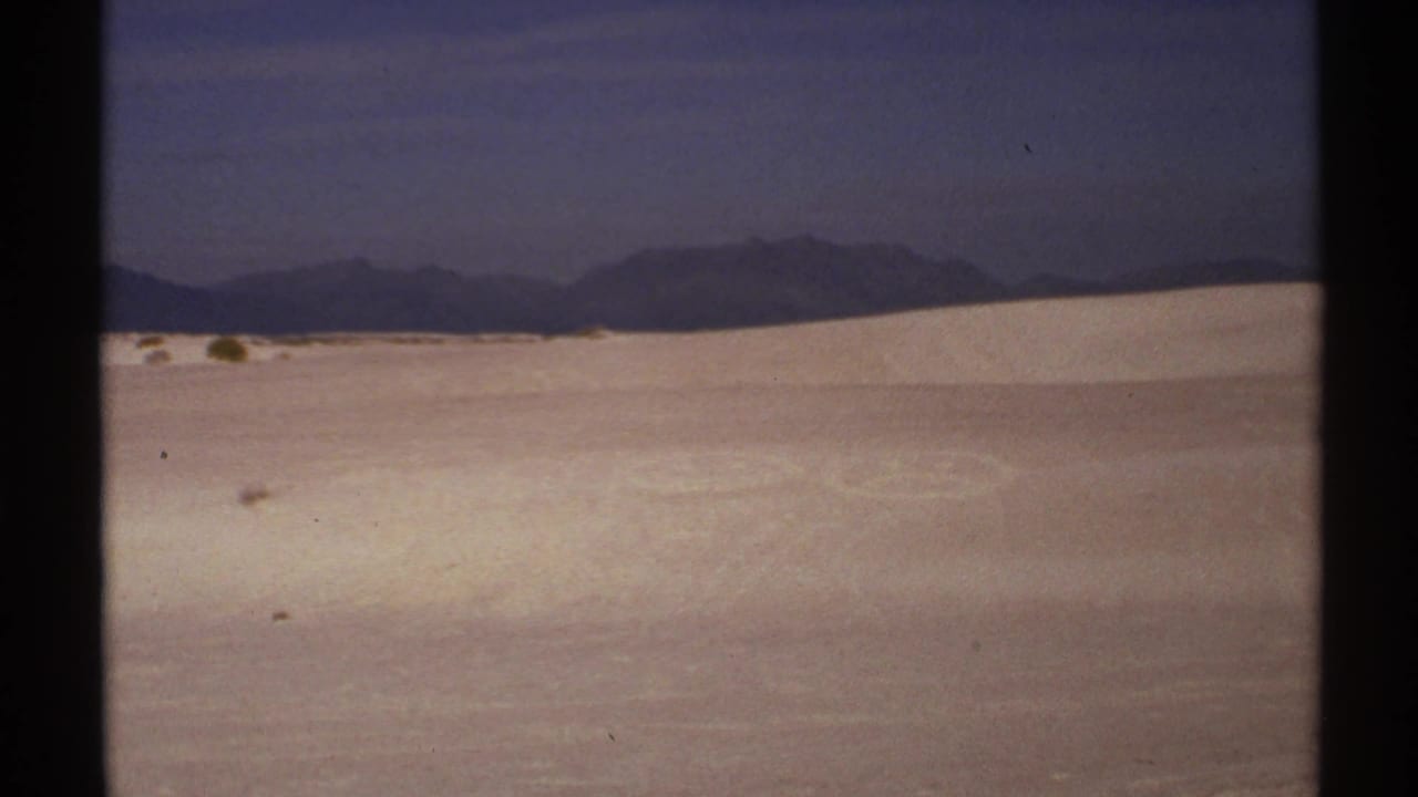 1972 New Mexico 8mm frame showing a wide desert sand landscape, dark sky, and distant mountain ridge.