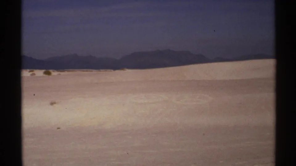 1972 New Mexico 8mm frame showing a wide desert sand landscape, dark sky, and distant mountain ridge.