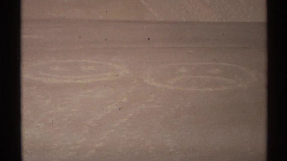 Close 8mm view of faint smiley and frowny face-like drawings scuffed into New Mexico sand.