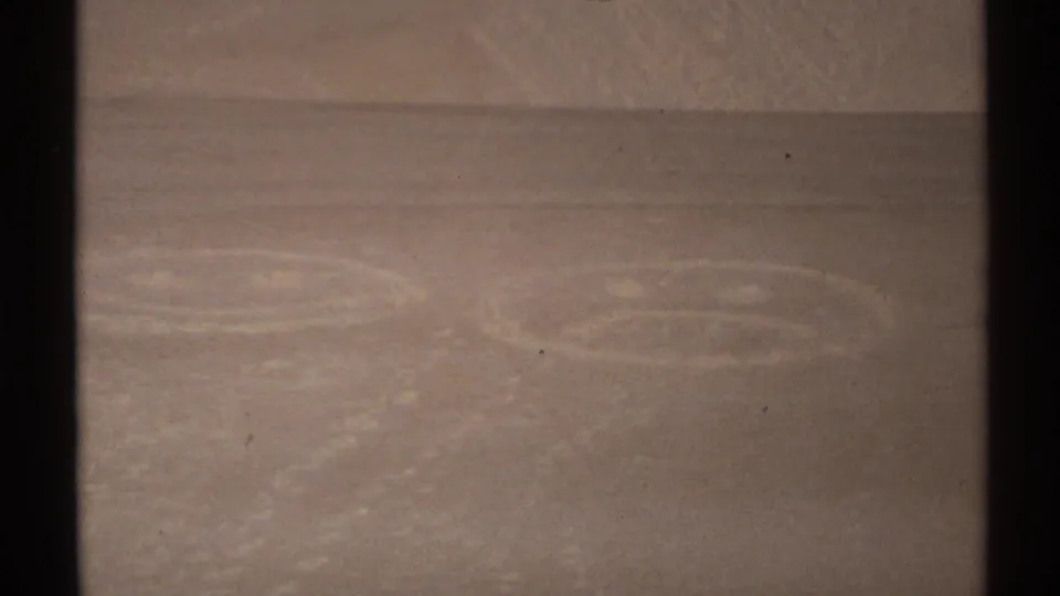 1972 New Mexico home-movie frame with trampled face-like sand drawings and footprints across a pale dune surface.