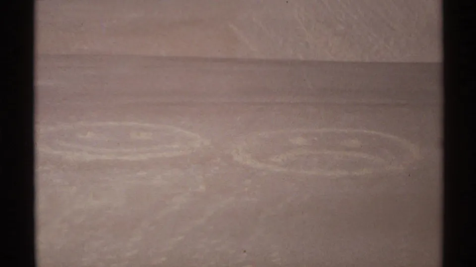 Close 8mm frame of two faint circular face designs in New Mexico sand, with trampling marks around them.