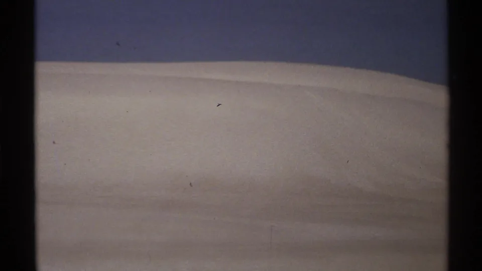 1972 New Mexico 8mm frame of a pale ridge-like outdoor surface beneath a dark blue sky, with scratches and dust in the scan.