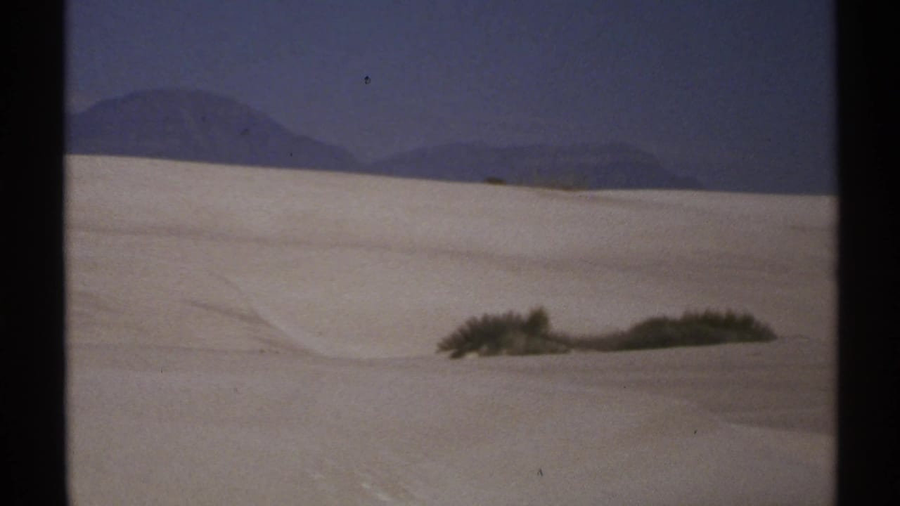 1972 New Mexico 8mm frame showing pale dunes, low desert shrubs, and a dark mountain horizon beneath a blue sky.
