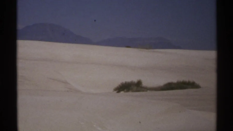 1972 New Mexico 8mm frame showing pale dunes, low desert shrubs, and a dark mountain horizon beneath a blue sky.