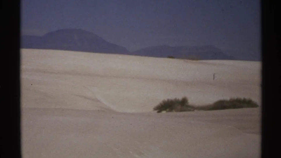1972 New Mexico 8mm frame with broad white dunes, a low shrub line, and hazy mountains beyond the sand.