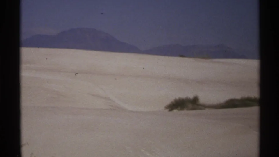 1972 New Mexico 8mm frame showing pale desert dunes, sparse shrubs, and a dark mountain range beneath open sky.