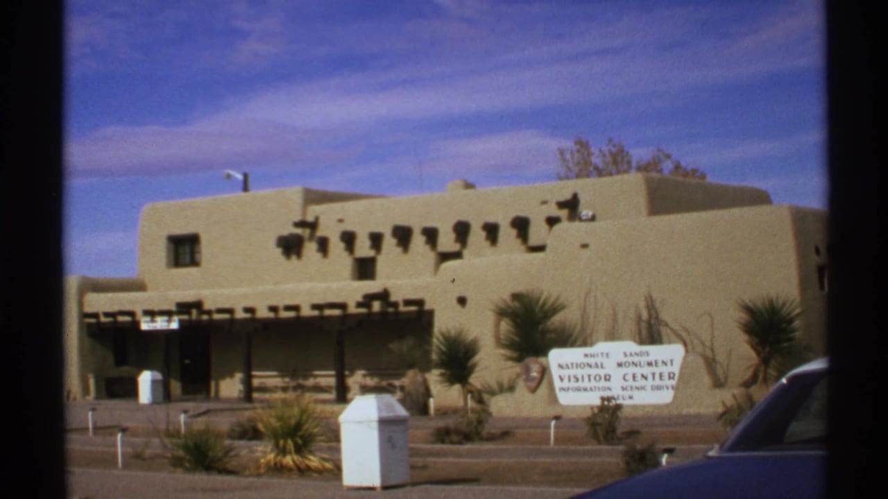 Visitor center facade, White Sands sign, yucca plants, blue sky, and dark car foreground.