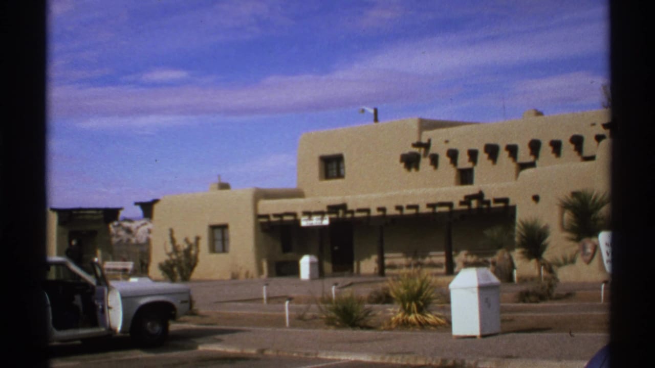 1972 New Mexico 8mm frame showing an adobe-style building, desert landscaping, and a white vehicle under a blue sky.
