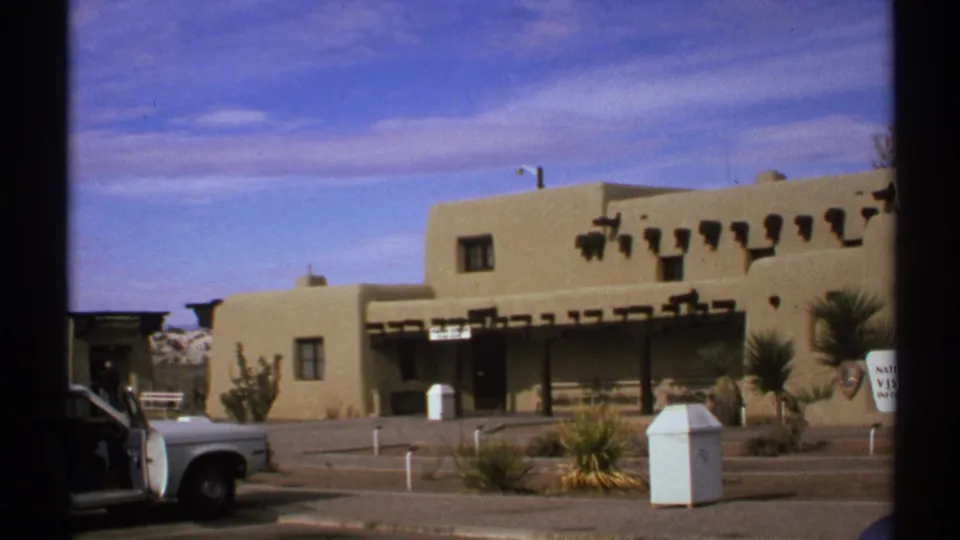 1972 New Mexico travel frame with adobe-style architecture, a shaded frontage, desert plants, and a white vehicle near the curb.