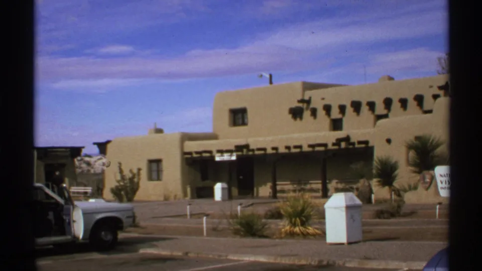 1972 New Mexico 8mm frame with adobe-style building details, roadside posts, desert plants, and a cropped white vehicle.