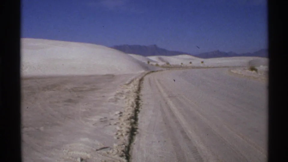 1972 New Mexico 8mm frame showing a dirt road through pale sand dunes with mountains in the distance.