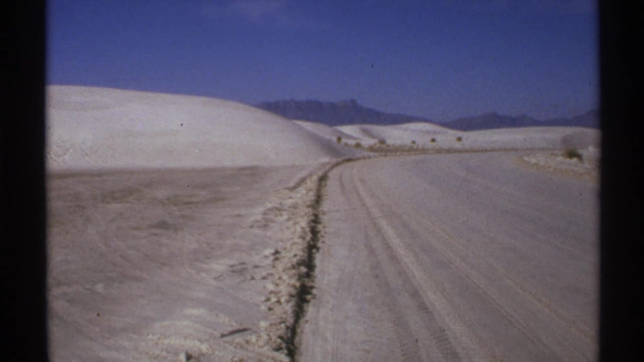 1972 New Mexico 8mm frame showing a dirt road curving through pale dune-like terrain toward distant mountains.